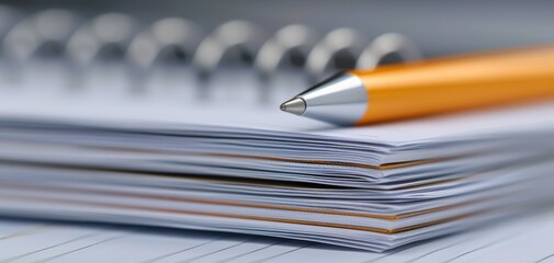 Stack of papers with an orange pen on top and a spiral notebook in the background.