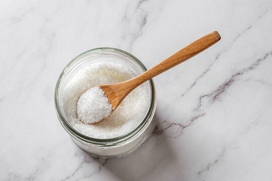 Full spoon in a jar of monosodium glutamate over kitchen marble counter. Flavor enhancer MSG white crystals in a glass container. E621 food additive concept.