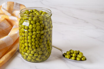 Open jar of canned green peas over marble countertop. Marinated sweet peas in a glass jar and metal spoon close-up. Vegetable protein, dietary fiber, salad ingredient.