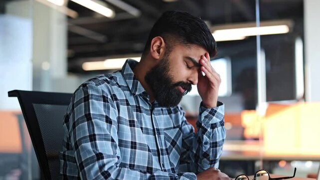 Tired businessman takes off glasses and rubs forehead from headache or fatigue. Frustrated man sitting at desk in modern office. Stressed worker experiencing burnout.