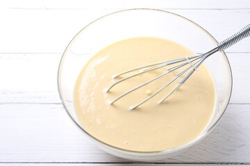 Whisk and bowl of dough on white wooden table, closeup