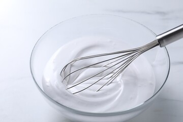 Whisk and whipped cream in bowl on white marble table, closeup