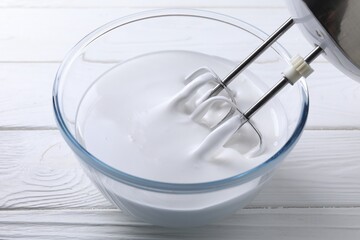 Mixing whipped cream with hand mixer in bowl on white wooden table, closeup