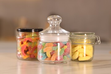Tasty gummy candies in glass jars on white marble table, closeup