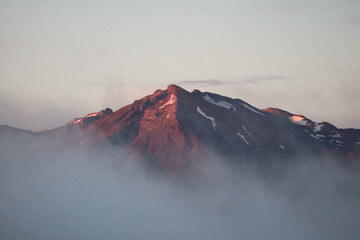A volcanic peak in New Zealand illuminated by the golden light of sunrise, with mist rolling through the valley below. A dramatic and breathtaking view.
