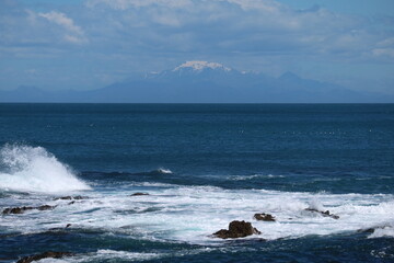 Powerful ocean waves crash against rugged rocks, with a snow-capped mountain in the distance. A breathtaking New Zealand seascape perfect for travel, adventure, and nature-themed visuals.