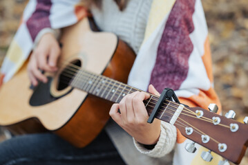 Close up of a young woman playing the guitar in autumn nature. Girl with guitar in hands, covered in warm plaid, playing the guitar outdoor in autumn forest, using capo