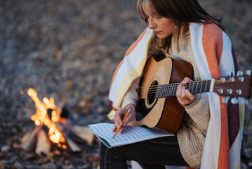 Girl composer playing the guitar on campsite near the bonfire. Young woman guitarist composing song outdoors in autumn evening park near bonfire, writing scores to her notebook