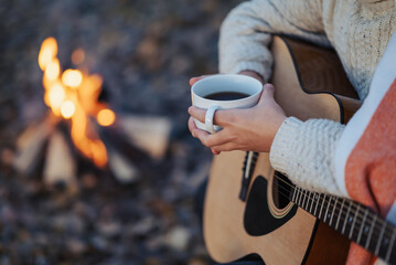 Girl with guitar and cup of coffee or tea near the campfire. Evening cosy shot of a young girl sitting near bonfire, playing the guitar, drinking hot coffee
