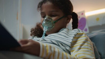 Medium close up of little sick Black girl in pyjamas breathing with oxygen mask lying on hospitable bed and drawing while receiving oxygen therapy