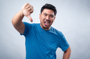 An Asian man in a blue t-shirt stands against a white background, giving a thumbs-down gesture with...