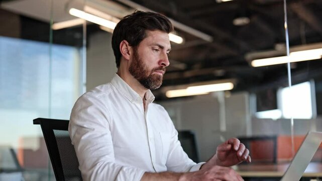 Focused businessman working on laptop in modern office. Bearded man in white shirt types on keyboard, concentrating on task.