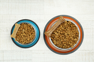 Dry pet food in feeding bowls and chew bones on floor, top view