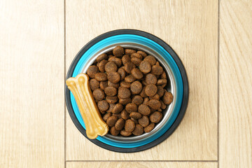 Dry pet food in feeding bowl and chew bone on wooden floor, top view