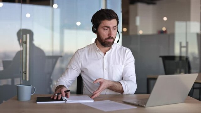 Confident businessman in headset having video call on laptop in modern office. Young male professional talking, discussing something with client or interviewing candidate.