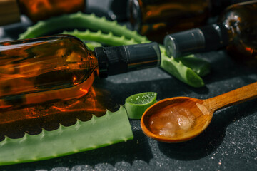 Close-up of fresh aloe vera slices and gel in a wooden spoon next to amber glass spray bottles on a...