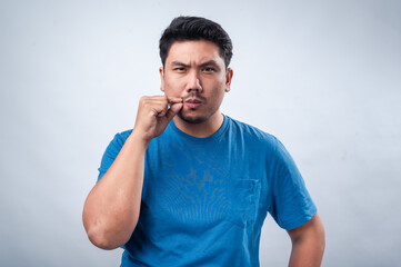 An Asian man in a blue t-shirt makes a silence gesture by placing his index finger and thumb near...