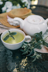 White teapot and cup of herbal tea with fresh mint leaves on a glass table. Soft fabric, straw hat, and white flowers in the background create a cozy outdoor setting for relaxation
