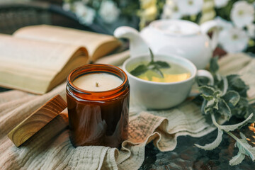 A cozy scene with a lit amber glass candle, a cup of herbal tea with mint, and an open book on a soft linen cloth. A white teapot and fresh greenery complete the tranquil atmosphere