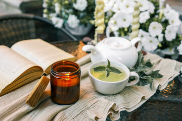 Cozy scene with a burning amber candle, herbal tea with mint, an open book, and a teapot on a linen-covered table. Warm ambiance with flowers and soft candlelight creating a relaxing atmosphere
