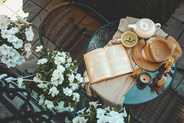 Top view of a cozy outdoor table with an open book, herbal tea, a straw hat, a candle, and blooming...