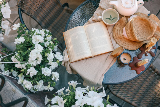 Top view of a cozy outdoor table with an open book, herbal tea, a straw hat, a candle, and blooming white flowers. Sunlight casts warm tones over the rustic and peaceful setting