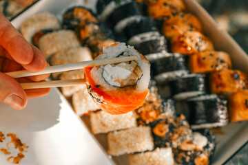 Assorted sushi rolls in a takeout box with nori, sesame seeds, grilled cheese, and spicy topping....