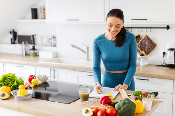 Slim lady in sportswear standing in kitchen and writing down healthy recipe or daily ration diet while cooking dinner at home, copy space