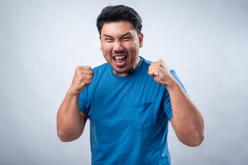 An Asian man in a blue t-shirt clenches his fists and shows an intense expression of excitement or triumph against a white background. His face conveys determination and strong emotions