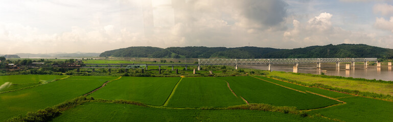 Scenic Green Fields With Flowing River And Steel Bridge Surrounded By Rolling Hills Under Partly Cloudy Sky