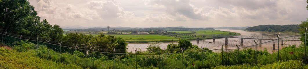 Expansive Panoramic View Of Green Fields And River With Bridges And Cable Cars Beyond A Barbed Wire...