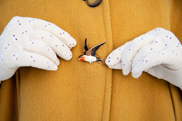 Vintage brooch in shape of flying swallow pinned to coat, close up. Lady's hands in white gloves. Retro accessories and outfit details. Yellow background.