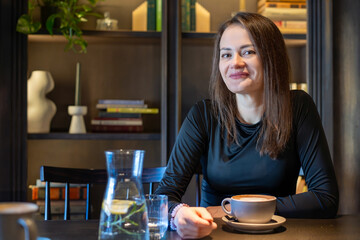 Smiling woman enjoying coffee at cafe. A smiling woman sits at a table in a stylish cafe, enjoying a cup of coffee with books and decor in the background.