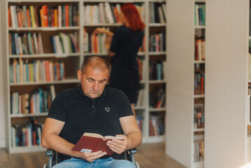 Man in wheelchair reading a book in library while librarian searching for book