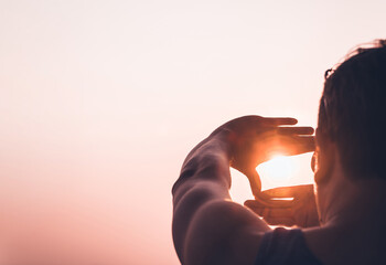 Silhouetted man framing the sun with his hands against a warm sunset sky, symbolizing vision,...