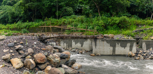 Unfinished bamboo bridge spanning rapid river with broken concrete supports amid lush green jungle...