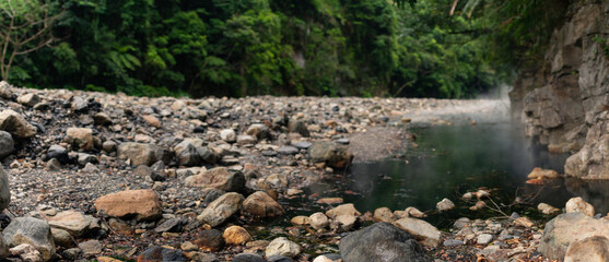 Thermal river with rising steam flowing through rocky landscape surrounded by dense tropical forest in wilderness