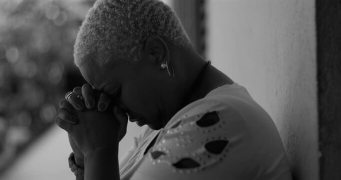 African American woman leaning against a wall, bowing her head in deep prayer, expressing emotional vulnerability and hope for strength amidst life&rsquo;s adversities