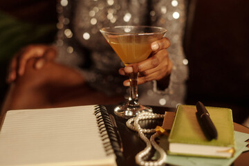 Person holding cocktail glass in elegant setting with pearl necklace and notebook on table....