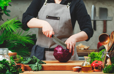 Chef cuts ​​red cabbage with nakiri knife. Cozy kitchen with wooden table, kitchenware, vegetables, herbs and ingredients for cooking. Healthy vegan eating, culinary, recipes, food blogging