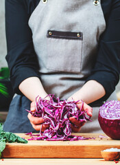 Chef holds ​​red cabbage sliced for salad in hands. Cozy kitchen with wooden table, kitchenware, vegetables, herbs and ingredients for cooking. Healthy vegan eating, culinary, recipes, food blogging 