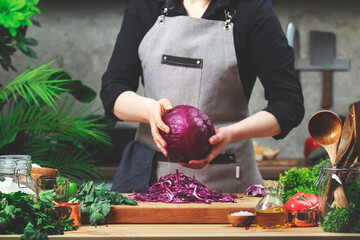 Chef a whole red cabbage in hands. Cozy kitchen with wooden table, kitchenware, vegetables, herbs and ingredients for cooking. Healthy vegan eating, culinary, recipes, food blogging comcept