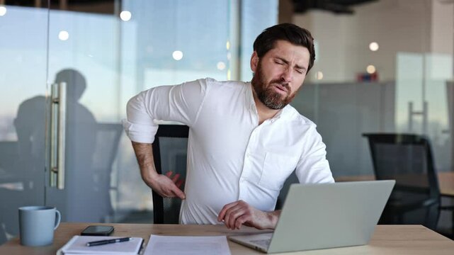 Exhausted businessman suffering from backache while working with laptop. Young male feeling pain after long time working at computer. Business office.