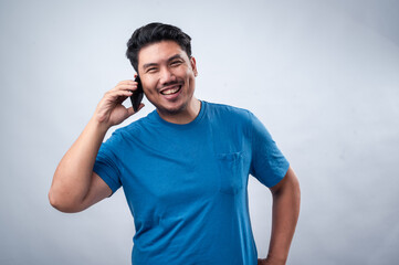 Fototapeta premium An Asian man in a blue t-shirt is talking on his smartphone with a cheerful expression. He is looking slightly upward while holding the phone to his ear. The background is plain white.