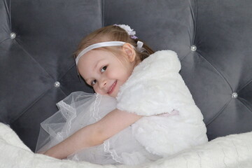little girl sitting in a white dress smiling to the camera. blond girl is hugging her legs with arms.