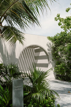 A minimalist white archway surrounded by lush tropical greenery, casting elegant shadows. A stone sign reads Summer Pavilion evoking a serene and modern architectural design in a tropical setting 