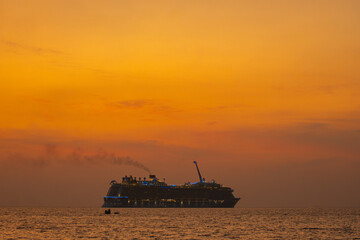 Obraz premium A large luxury cruise ship sails across the ocean under a vibrant orange sunset sky. The ship's lights glow against the warm hues of the evening, creating a serene and picturesque maritime scene.