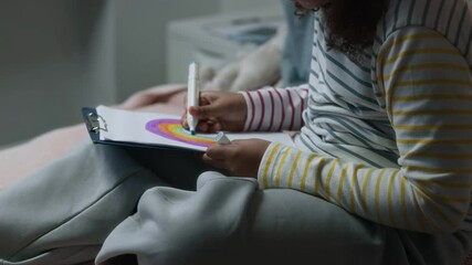 Little talented African American girl wearing comfortable pyjamas sitting on hospitable bed and drawing beautiful colorful rainbow on paper using marker