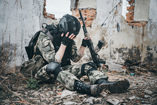 A soldier in uniform with a machine gun sits on the ruins of a destroyed building and covers his face with his hand. Post-traumatic stress disorder concept