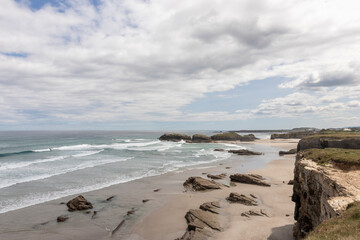 A scenic coastal landscape featuring a sandy beach, rugged rock formations, and ocean waves under a partly cloudy sky. A peaceful and natural seaside view